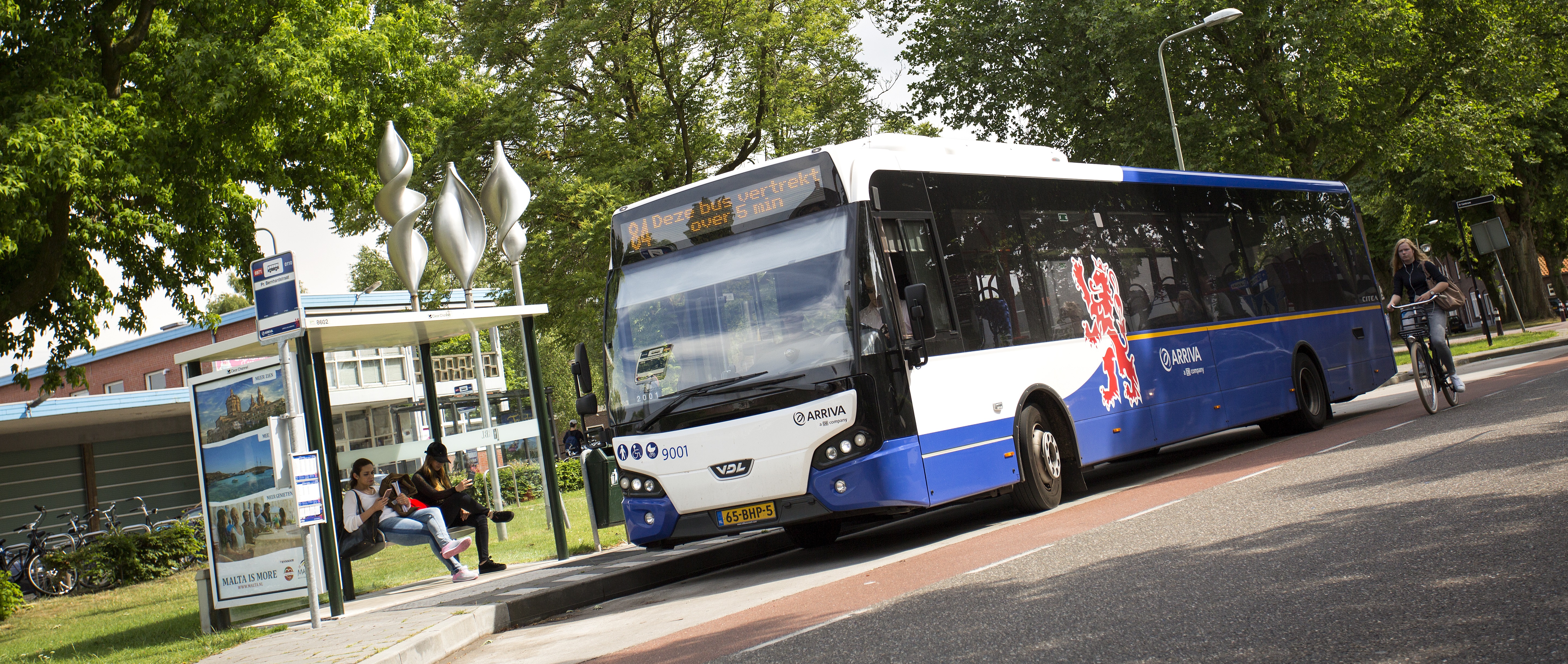 Bushalte in Venray met wachtende reizigers en een passerende Arriva-bus, in een groene stedelijke omgeving – beeld van de bereikbaarheid in de regio.