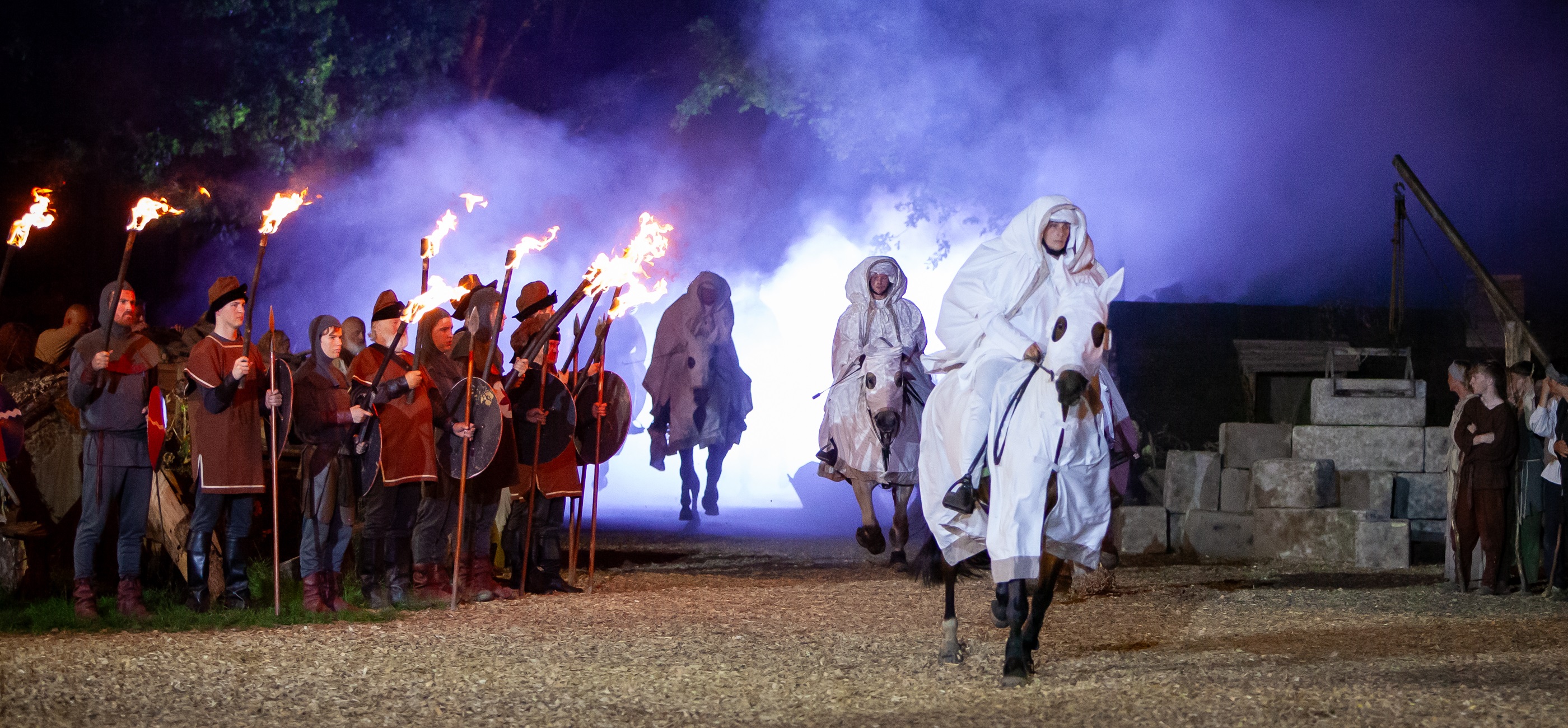 Historische scène van het Draaksteken Beesel met ridders in middeleeuwse kleding, fakkels en ruiters op witte paarden in een sfeervol verlicht decor.
