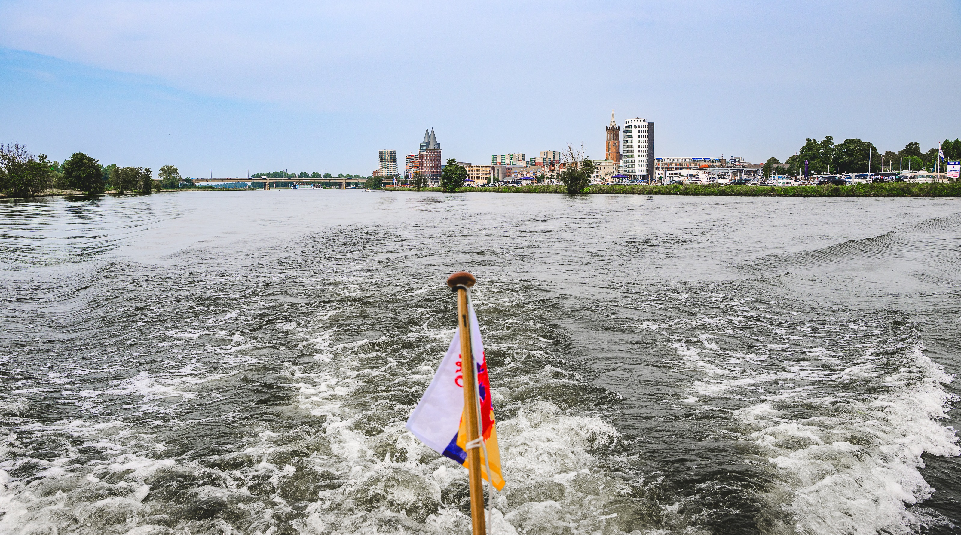 Uitzicht vanaf een boot op de Maasplassen bij Roermond, met in de verte de skyline van de stad en de Limburgse vlag op de voorgrond.