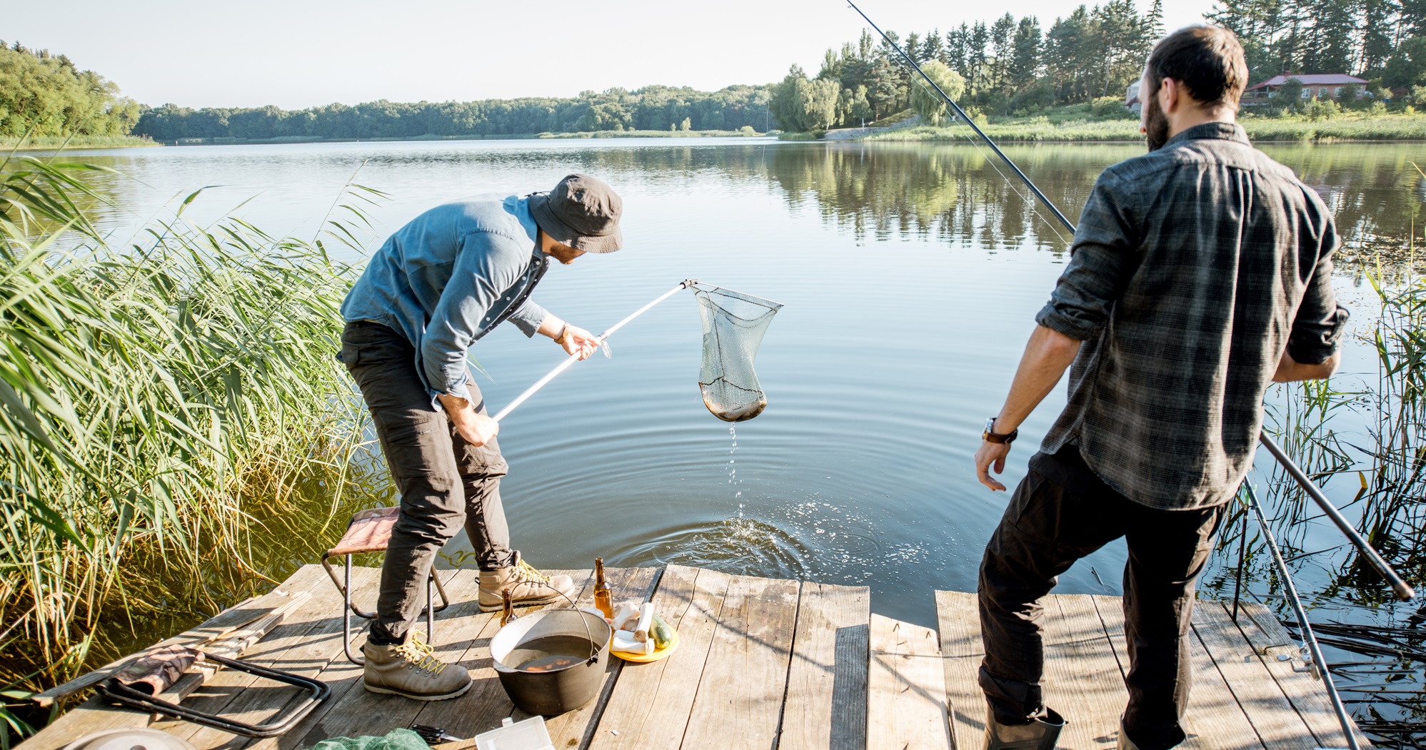 Twee mannen aan het vissen in Limburg vanaf een houten steiger aan een rustige visvijver, omringd door riet en bos.