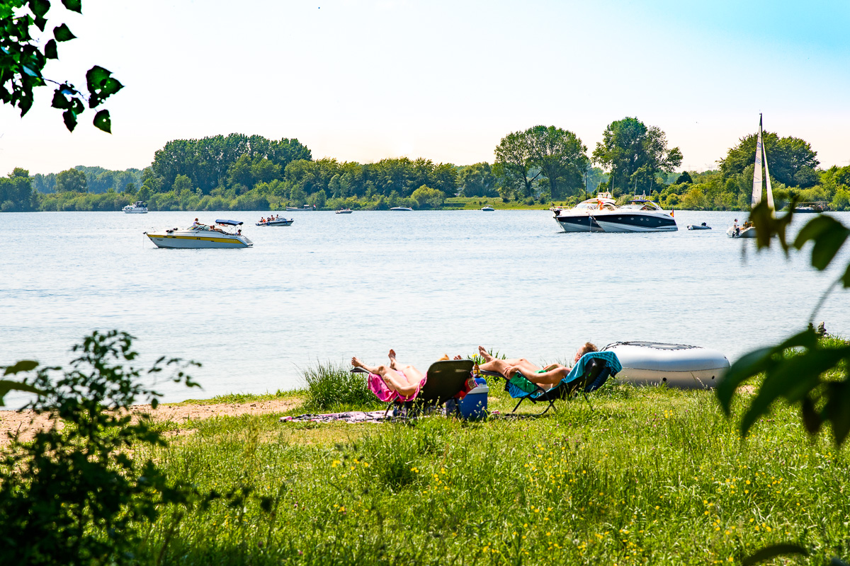 Mensen ontspannen op ligstoelen aan de oever van de Maasplassen, met op de achtergrond verschillende boten op het water op een zonnige dag.