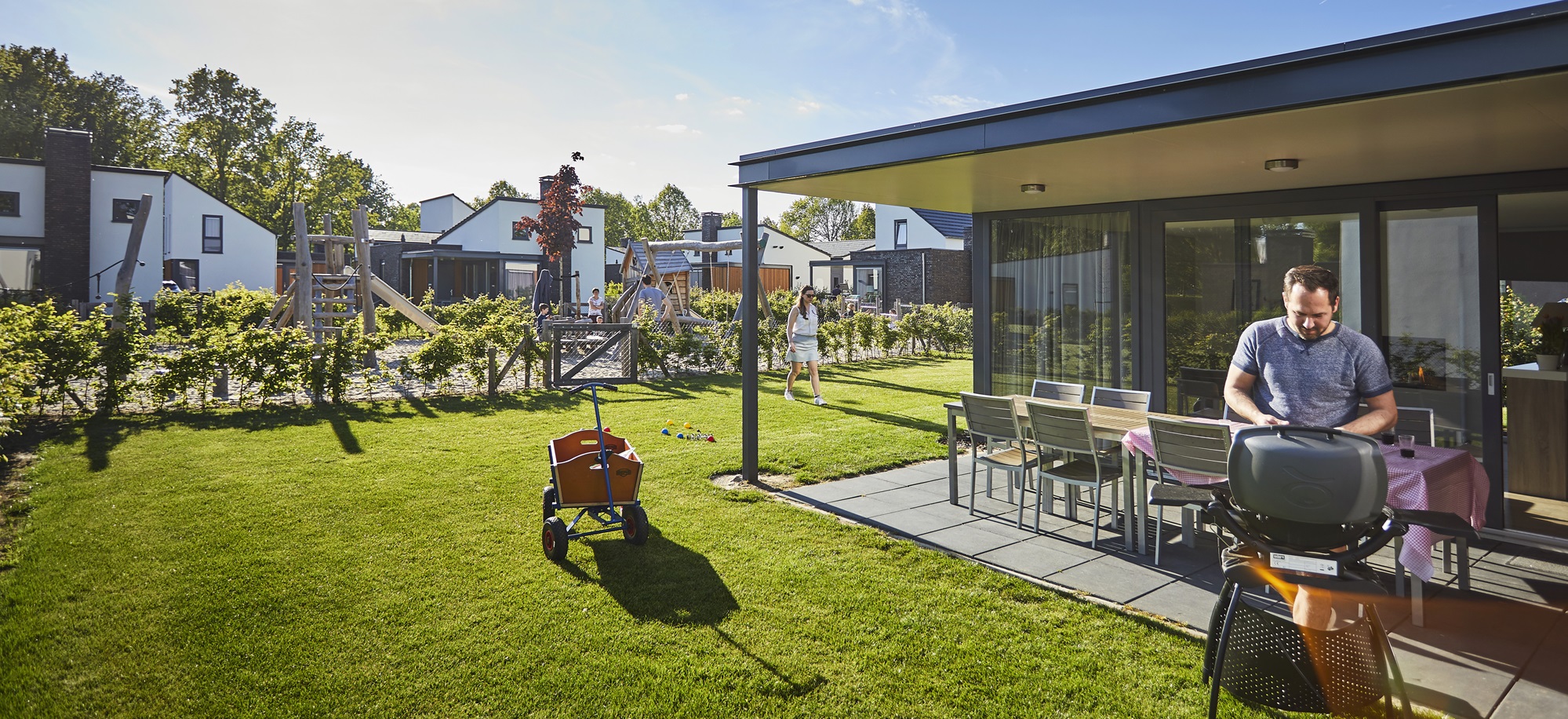 Gezin geniet van een zonnige middag in de tuin van een vakantiebungalow op recreatiepark de Leistert, met speeltuin op de achtergrond en een man die aan het barbecueën is.