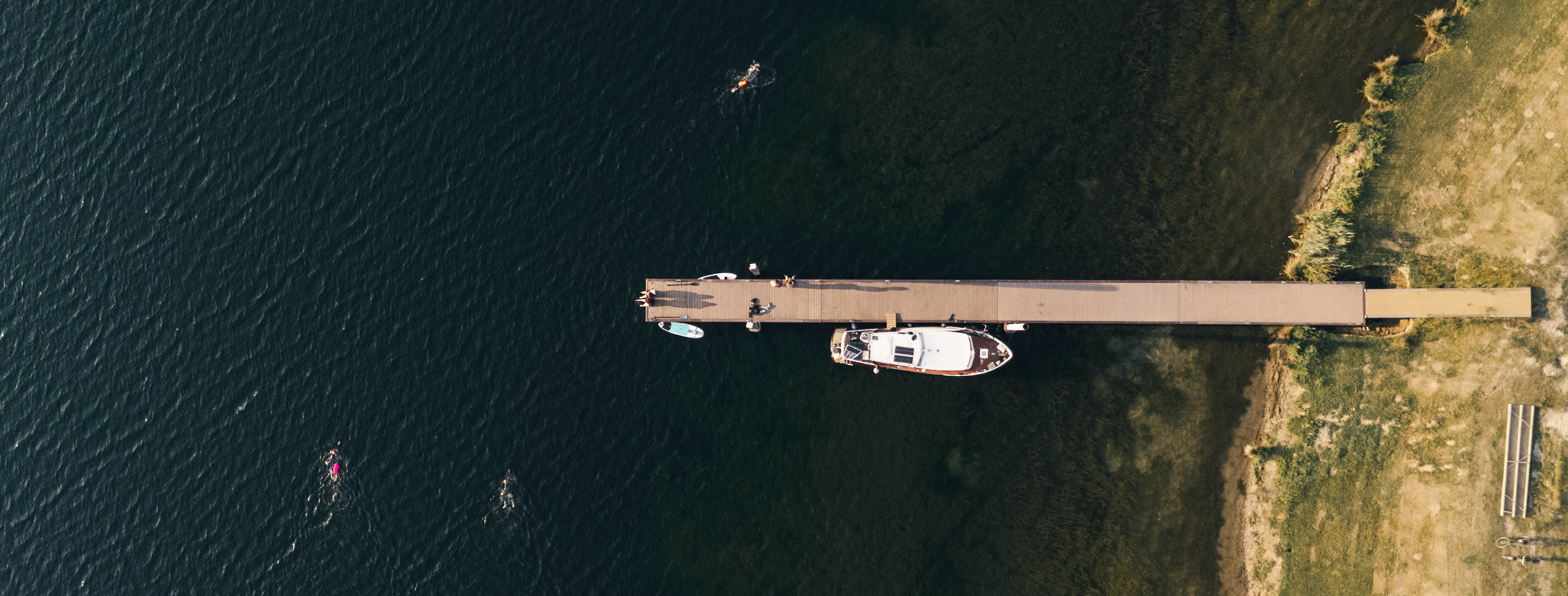 Luchtfoto van een lange houten steiger die vanaf de oever het water inloopt, met een aangemeerde boot en een SUP-board. In het water rondom de steiger zwemmen meerdere mensen.