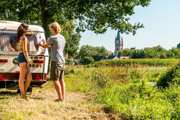 Zwei Personen in einem Retro-Wohnwagen am Rande eines Feldes, mit Blick auf die weiße Stadt Thorn und einen Kirchturm unter einem klaren Sommerhimmel.