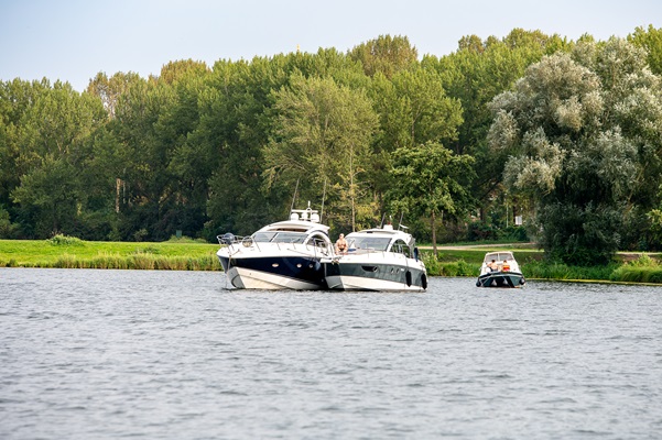 Drei Motorboote liegen auf der ruhigen Maas mit einem grünen Ufer voller Bäume im Hintergrund.