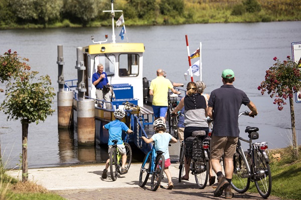 Radfahrer, darunter Kinder und Erwachsene, besteigen an einem sonnigen Tag mit ihren Fahrrädern eine Fähre über die Maas in Blitterswijck.