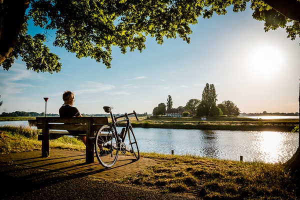 Wielrenner rust uit op een bankje aan de Maas en geniet van de zonsondergang in Stevensweert
