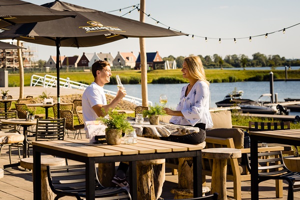 Twee mensen proosten op een zonnig terras aan het water met uitzicht op boten en moderne huizen in de verte.
