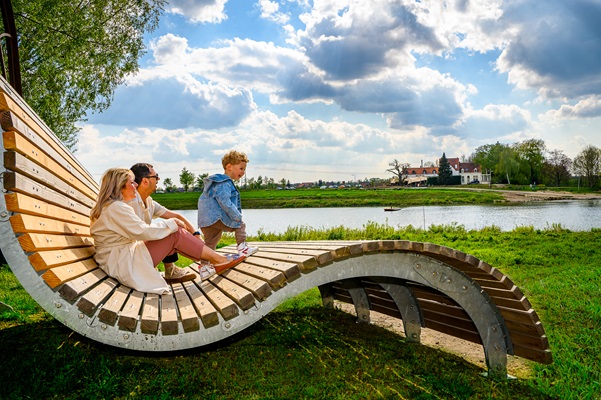 Een gezin geniet van het uitzicht vanaf een kunstzinnig houten bankje aan de Maas met het klooster van Steyl in de verte.
