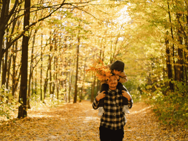 Ein Mann geht durch den Wald mit einem Kind auf seinen Schultern, das Blätter in den Händen hält. Der Mann und das Kind schauen sich an.
