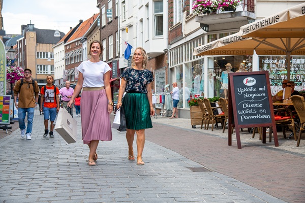 Zwei Frauen gehen mit Einkaufstaschen durch die Innenstadt von Roermond, vorbei an Geschäften und einer Terrasse mit einer Menütafel.