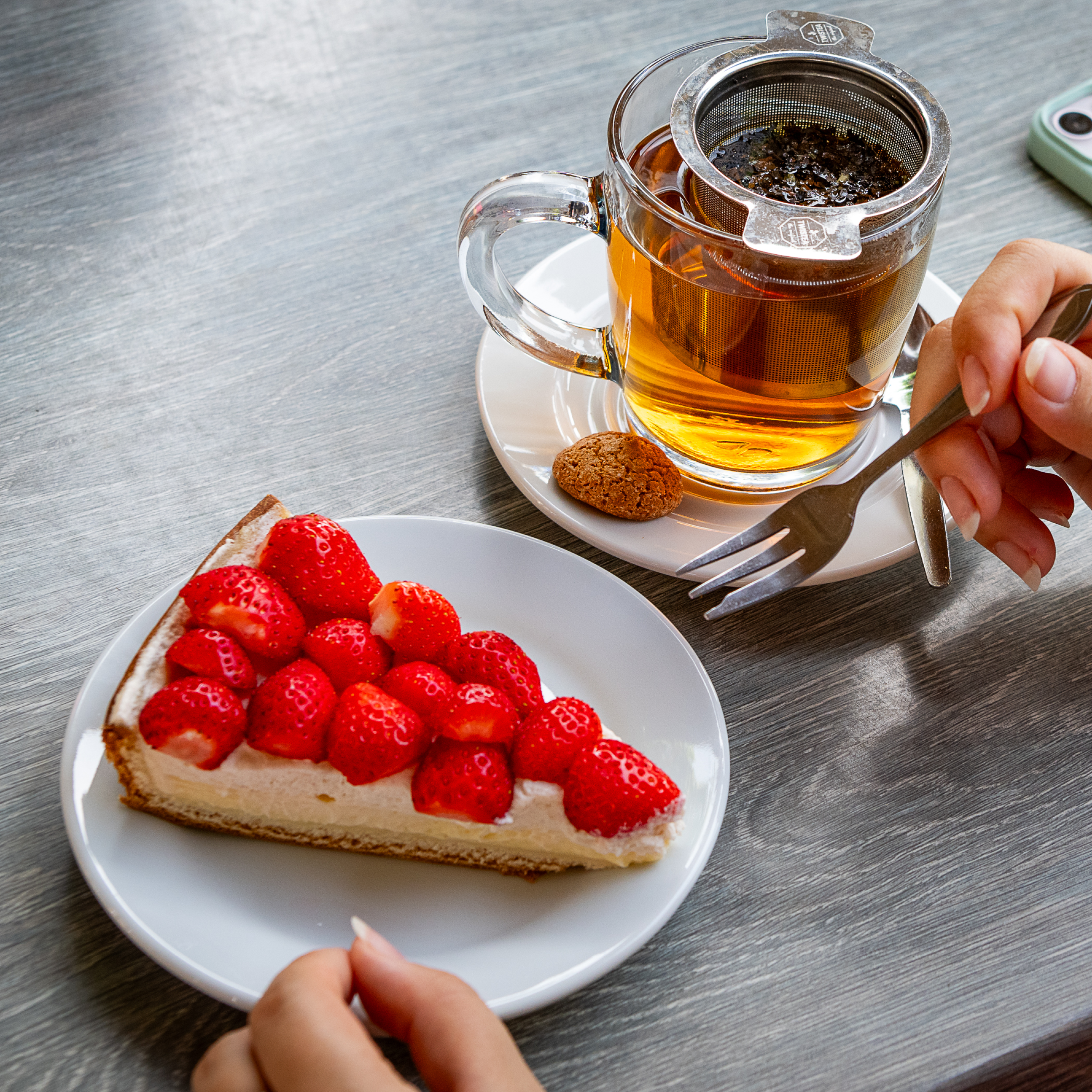 “Een punt aardbeientaart op een wit bord, met daarnaast een glas thee met een theezeefje. Iemand houdt een vork vast en staat op het punt een hap te nemen.”
