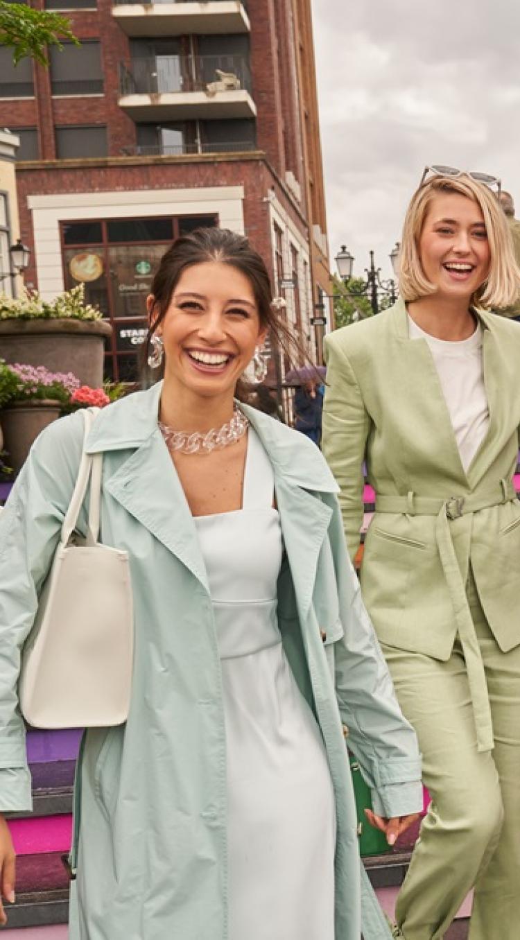 Vier vrouwen lopen lachend de kleurrijke trappen af bij Designer Outlet Roermond, gekleed in modieuze outfits, met winkels en bezoekers op de achtergrond.