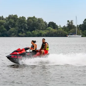 Twee personen rijden op een waterscooter over de Maasplassen, met op de achtergrond een zeilboot en groene oever. Actieve watersport op het water in Limburg.