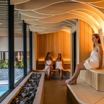 Drie vrouwen ontspannen in een moderne houten sauna met sfeervolle verlichting en uitzicht op het zwembad.