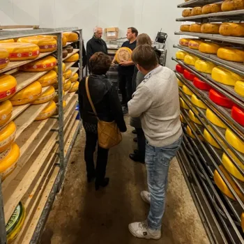 Bezoekers krijgen een rondleiding in een kaasmakerij, tussen rekken vol kazen. Een medewerker toont een kaaswiel en geeft uitleg.