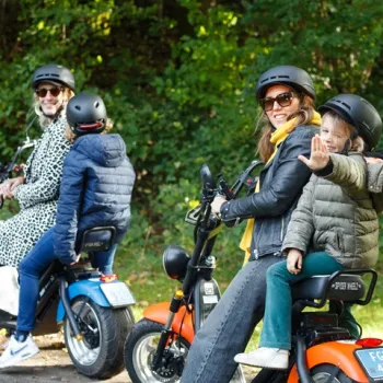 Vrouwen en kinderen op elektrische scooters tijdens een groepsuitje in een groene omgeving. Eén kind zwaait vrolijk naar de camera.