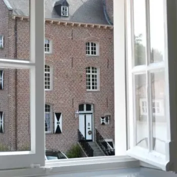 Uitzicht vanuit een open raam op de historische gevel van Kasteel Aldenghoor, met rood-witte luiken en een monumentale ingang.